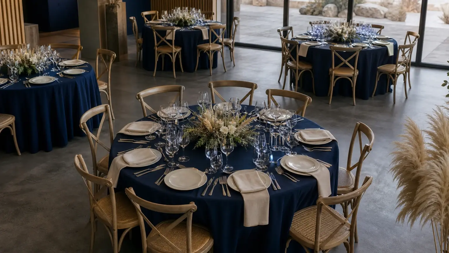 Salle de réception avec plusieurs tables rondes dressées, nappées de bleu, entourées de chaises et décorées de compositions florales.